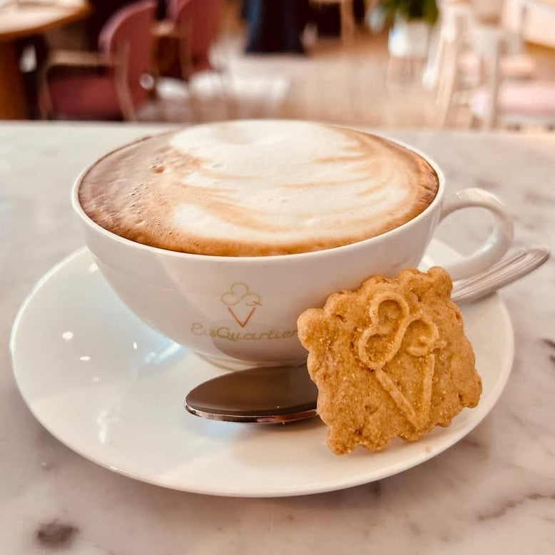 Gastro-Logokeks mit Eiswaffel-Motiv an einer Tasse Cappuccino, serviert auf einem Marmortisch im Café-Ambiente