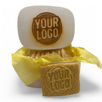 Holzkästchen mit gelasertem Logo im Deckel, gefüllt mit 8 Logokeksen in gelbem Seidenpapier – ein Keks zur Ansicht vorn platziert